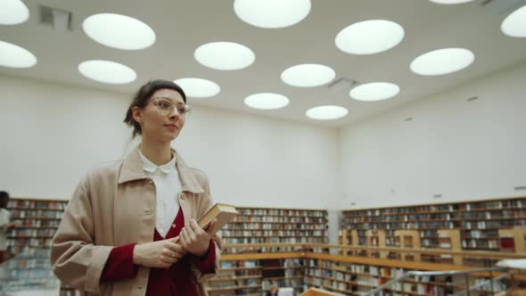 Woman Holding Book and Walking in Library