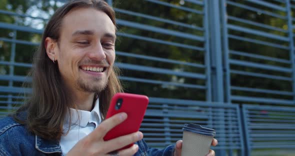 Close Up View of Joyful Long Haired Guy Holding Paper Coffee Cup and Laughing While Reading Funny