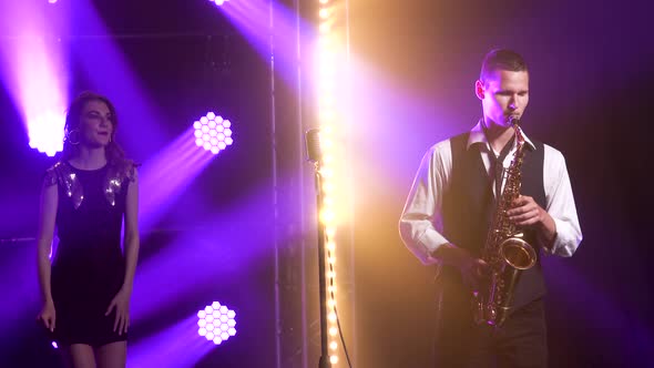 A Young Stylish Guy Plays the Saxophone on Stage and a Girl in a Black Dress with Sequins Is Dancing