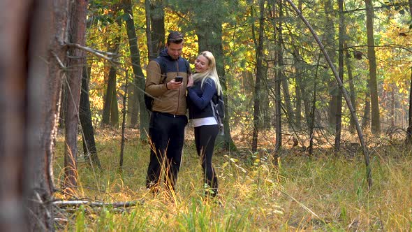 A Hiking Couple Stands in the Middle of a Meadow in a Forest and Takes Selfies with a Smartphone