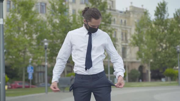 Portrait of Sad Man in Covid-19 Face Mask Showing Empty Pockets. Unemployed Caucasian Young