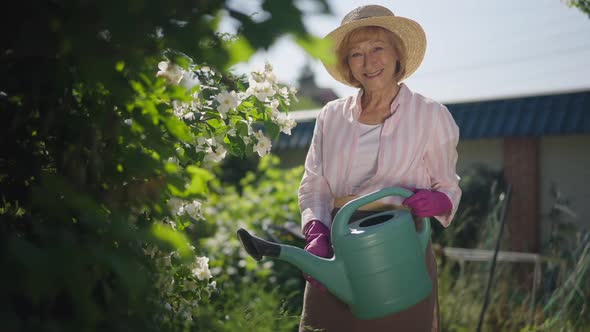 Portrait of Smiling Senior Woman with Watering Can Looking at Camera and Watering Plants in Garden