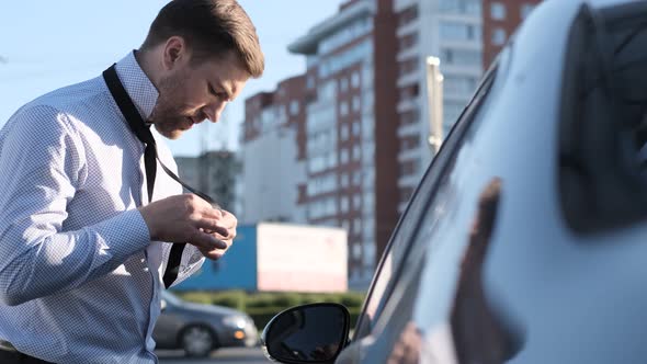 Young Businessman Man Puts on a Tie on the Street Looking Out the Window of His Car