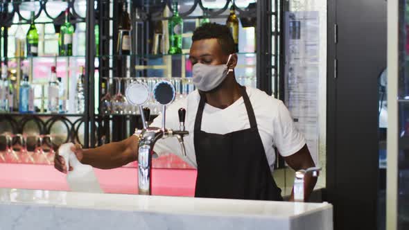 African american male barista wearing face mask disinfecting bar surface