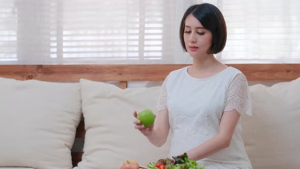 Beautiful Asian pregnant woman sit on sofa in living room then pick apple up and hold