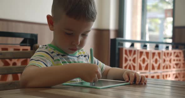 A Portrait of a Child Sits at a Table at Home and Draws on His Tablet with a Special Pen a