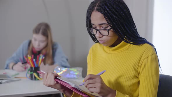 Smart African American Girl Writing in Notebook, Fixing Eyeglasses, Looking at Camera and Smiling