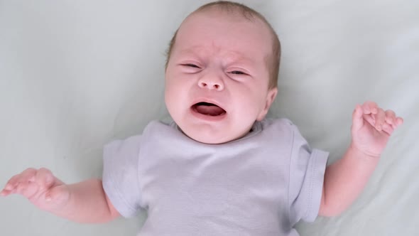 Dissatisfied Crying Upset Sad Newborn Baby Boy Lying in Nursery in Crib with Blue Clothes on Bed in