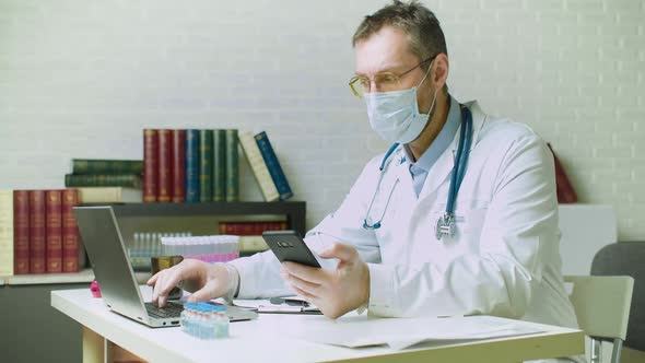 Middleaged Doctor Checks Mail From a Patient on His Smartphone and Enters the Data Into His Laptop