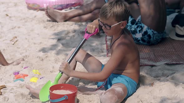 Blond Boy in Glasses Plays with Kids Spade and Sows Sand