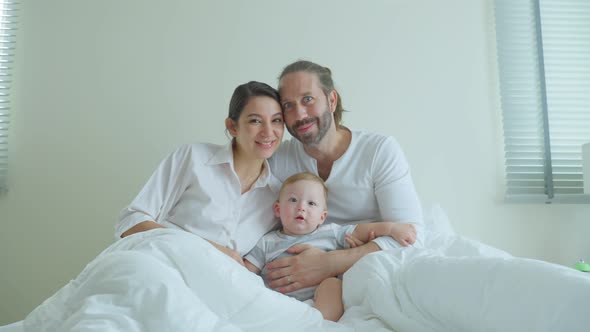 Portrait of Caucasian happy family, father and mother sit on bed with little baby boy on bed in room