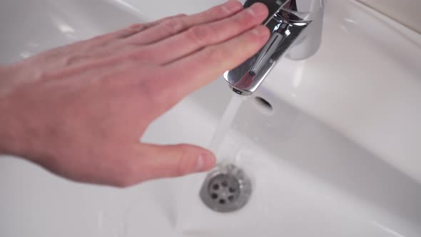 A man's hand closes a faucet in a white bathroom sink and tap water stops pouring