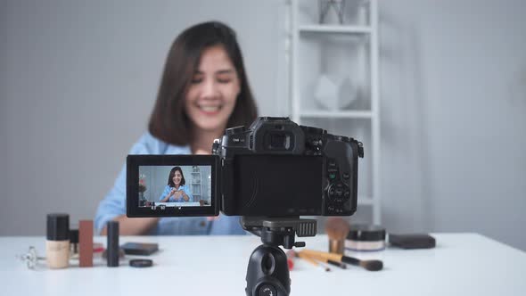 Asian woman or beauty blogger with brush and camera recording video and waving hand at home
