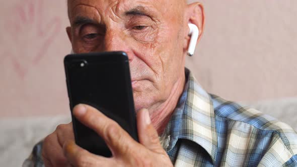 Portrait Caucasian pensioner 70-79 years old in modern wireless headphones uses a smartphone. The pe
