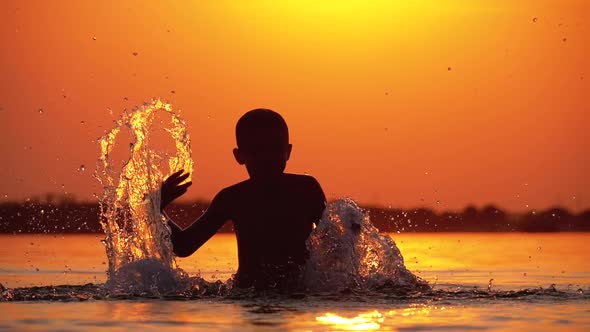 Silhouette of Boy at Sunset Raises Hands and Creating Splashes of Water. Slow Motion