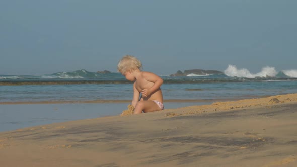 Girl Sits on Beach Playing with Sand Against Ocean Waves