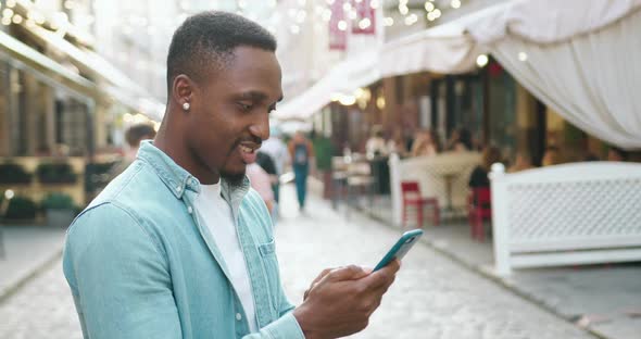 Black-Skinned Stylish Guy Standing on the Urban Street with Outdoors Cafes and Uses His Smartphone