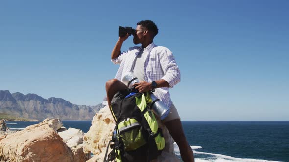 African american man hiking using binocilars sitting on rock by the coast