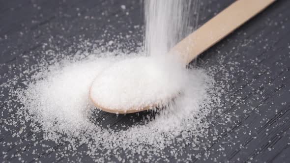 Crystals of white granulated sugar fall into a wooden spoon 