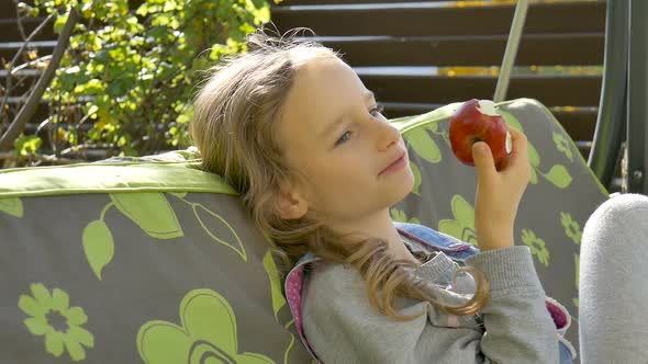 Little Child Blond Girl is Eating Red Apple on a Swing Outdoor During Summer Sunny Day on Playground