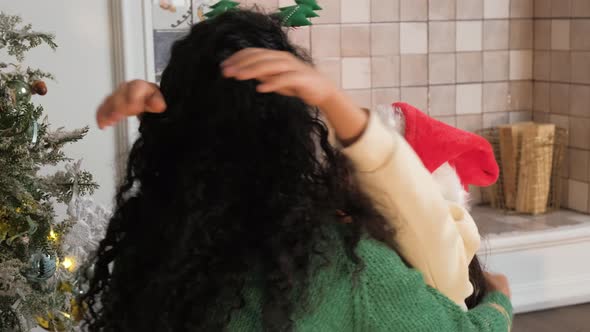 Little Girl in a Christmas Hat Hugs Her Mother Against the Background of a Festive Fir Tree and