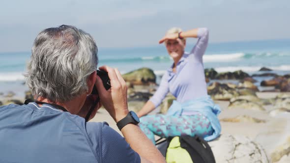 Senior hiker man taking pictures of senior hiker woman using digital camera on the beach.