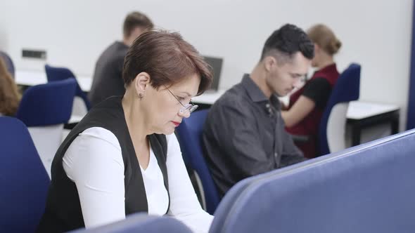 Side View of Focused Middle Aged Woman in Eyeglasses Working in Office with Blurred Coworkers at