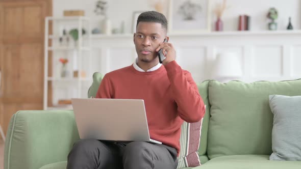 African Man with Laptop Talking on Phone on Sofa