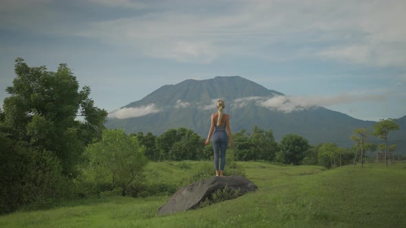 Spiritual yoga woman steps onto boulder raising arms into upward salute pose towards mount Agung