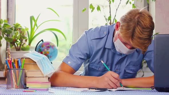 Teenage Man in a Blue Medical Mask of Caucasian Ethnicity Sits at His Desk at His Desk 