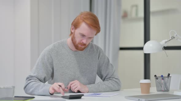 Redhead Man Using Calculator While Writing on Paper 