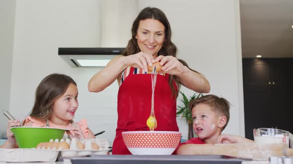 Happy caucasian mother, daughter and son breaking eggs, baking together in kitchen