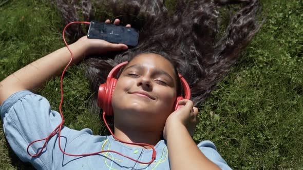 Attractive Girl with Long Hair Lies on the Grass in the Park and Listens to Music in Red Headphones