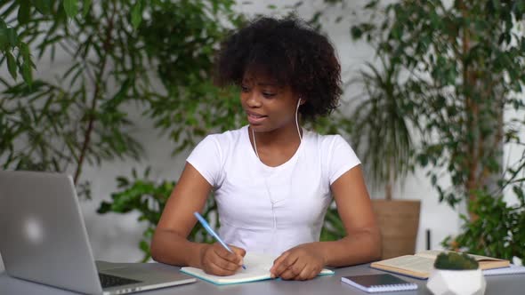 Smiling Mixed Race Female Student Writing Notes in Copybook Looking at Computer Screen Studying