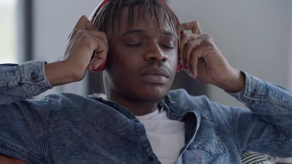 Portrait of Young African American Man Putting on Headphones and Leaning on Comfortable Couch in