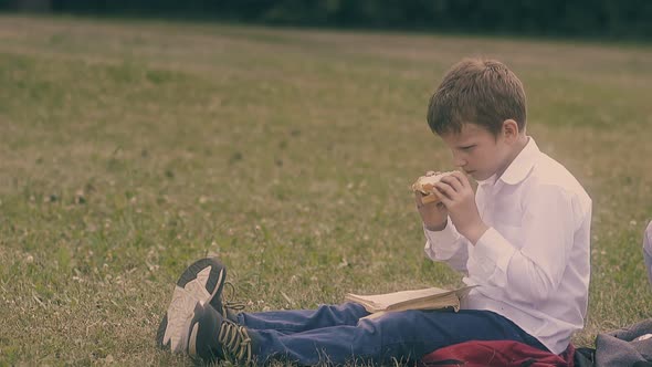 Boy with Fair Hair Eats Sandwich Sitting on Green Grass