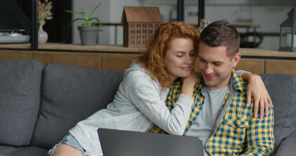 Young Sweet Couple Is Sitting on a Sofa, Browsing in Laptop