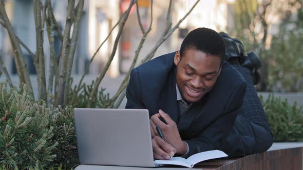 Smiling African American Student Trainee Lying on Stomach Using Laptop Making Notes on Notepad