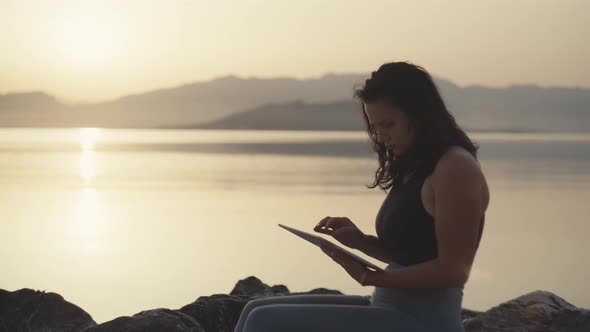 Girl Sitting with a Tablet By the Sea After Jogging at Sunrise