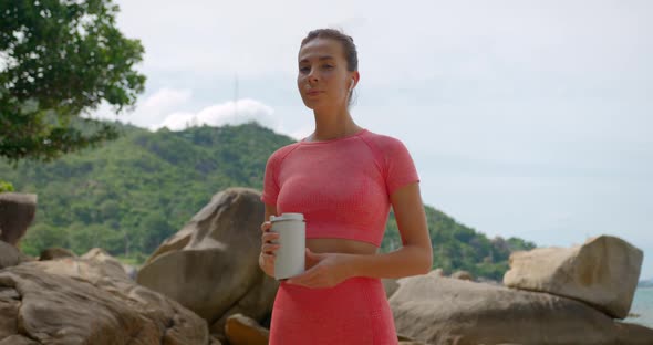 Young Woman in Pink Yoga Suit with Bottle of Water in Hand Standing Among Stones on Tropical Beach