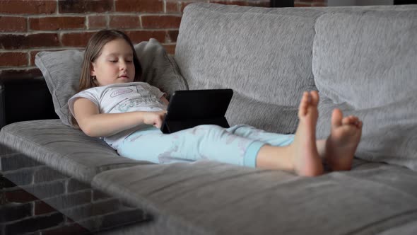 A little girl lying at home on the couch and looks into the black tablet. The child plays