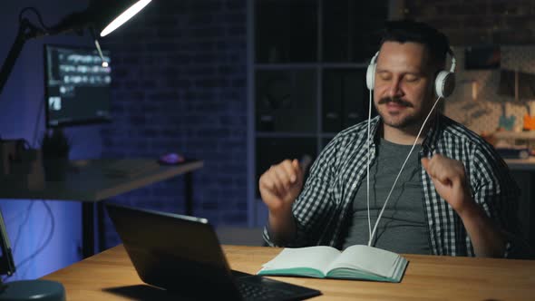 Joyful Man Enjoying Music in Headphones Busy with Laptop Dancing in Dark Office