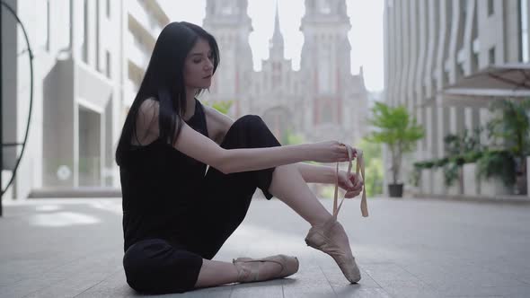 Serious Elegant Ballerina Tying Pointe Shoes Sitting on Asphalt with Ancient Buildings 