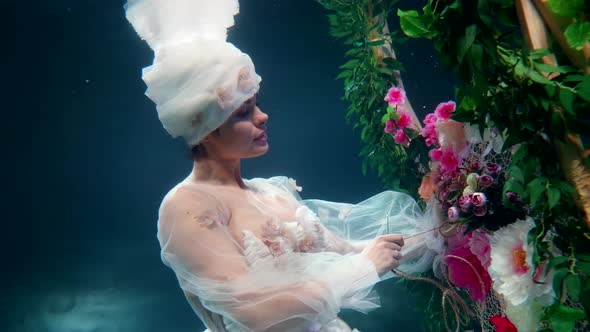 Floral Fairy Inside Depth of Magical Sea Lady in White Gown is Sewing Flowers to Fence Underwater