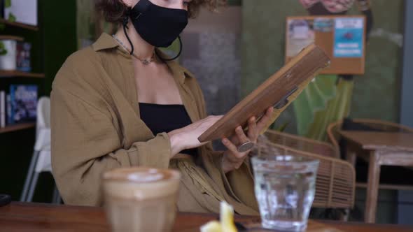 Side View of Cappuccino Served in a Glass on a Wooden Tray and Young Woman in a Face Mask Checking