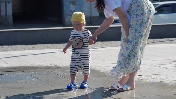 Happy Kid with His Mother Having Fun in Fountain of Public Park at Sunny Summer Day