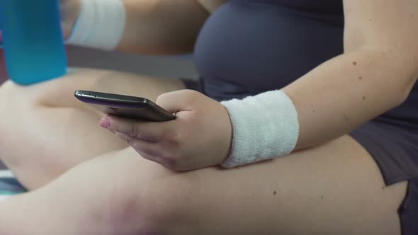 Young Woman in Sportswear Sitting on Floor, Scrolling Cellphone, Drinking Water