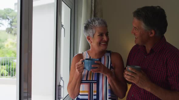 Caucasian senior couple smiling and drinking coffee together while looking out of the window at home