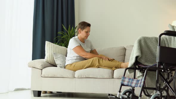 Senior Woman Massaging Her Legs While Sitting on the Couch Near the Wheelchair