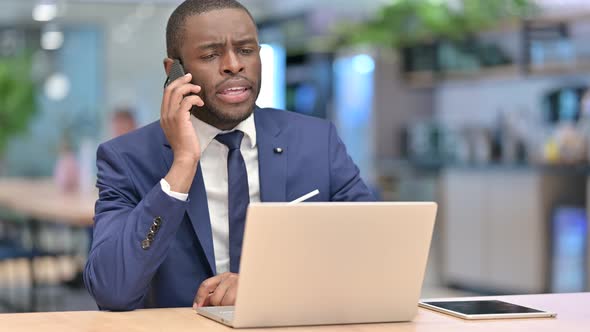 African Businessman with Laptop Talking on Smartphone in Office 